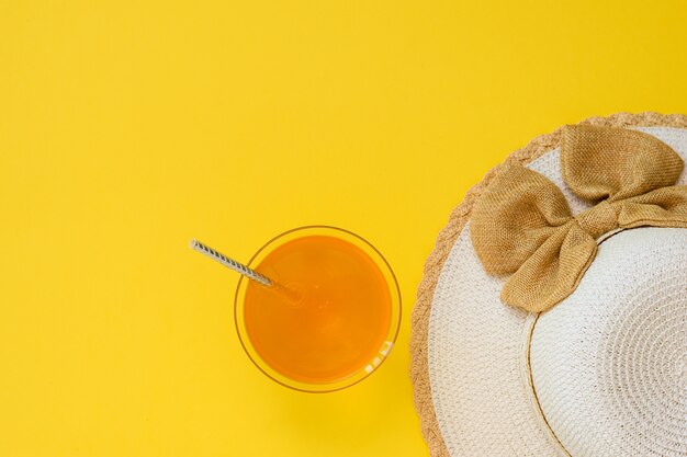 Sombrero y bebida de naranja en un tazón con paja cóctel sobre fondo amarillo. El concepto de vacaciones de verano. Endecha plana. La vista desde arriba.