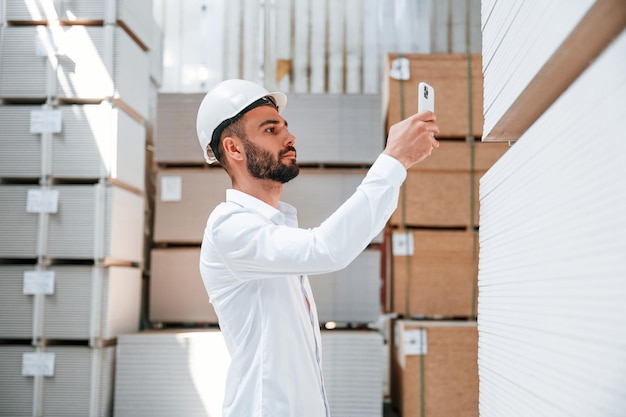 Con sombrero duro blanco, el trabajador de almacenamiento está en el almacén con un montón de productos