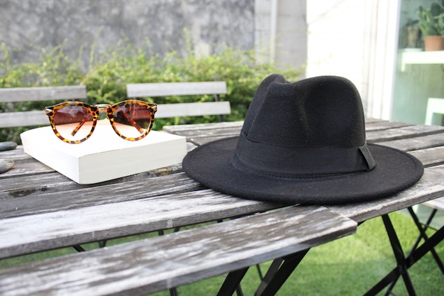 Sombrero Fedora, gafas de sol y libro sobre mesa de madera al aire libre, día de enfriamiento.