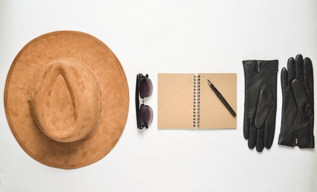 Foto sombrero de fieltro beige, cuaderno con bolígrafo, gafas de sol, guantes sobre un fondo blanco.