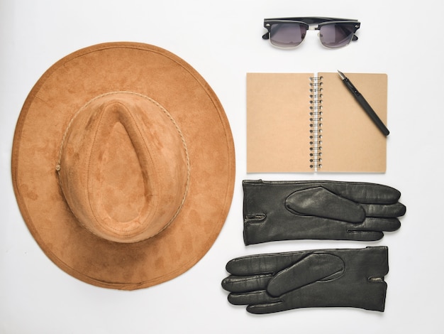 Sombrero de fieltro beige, cuaderno con bolígrafo, gafas de sol, guantes sobre un fondo blanco.