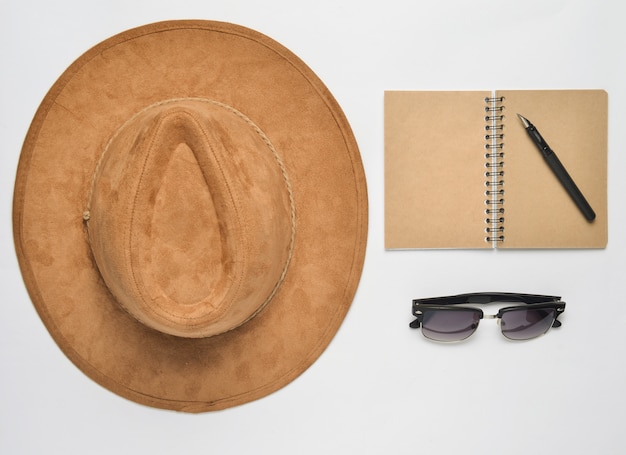 Foto sombrero de fieltro beige, cuaderno con bolígrafo, gafas de sol sobre un fondo blanco.