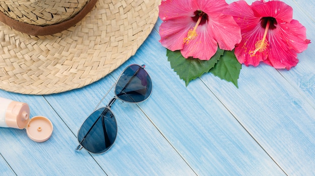 Sombrero, gafas de sol, bloqueador solar y rosa china en una mesa de madera azul.