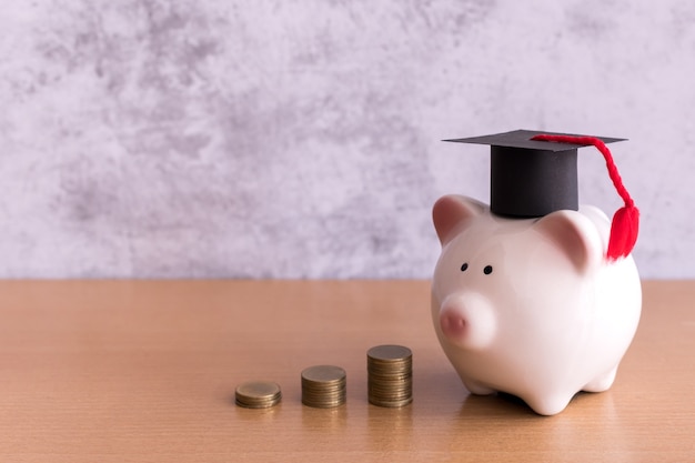 Sombrero de graduación en hucha con pila de monedas dinero en la mesa, ahorrando dinero para el concepto de educación