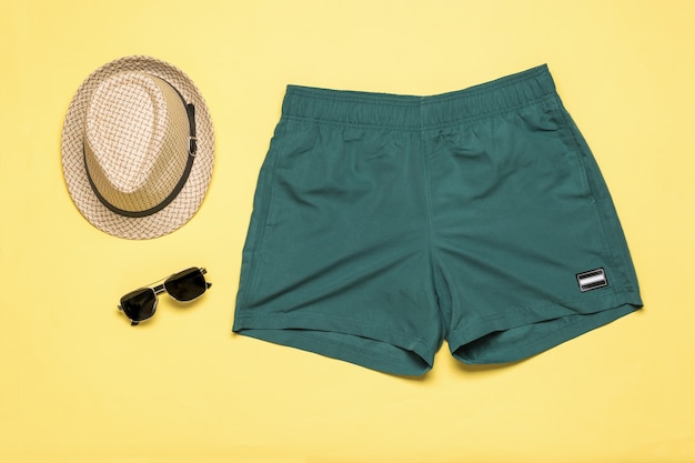 Sombrero de hombre con gafas y shorts de playa sobre un fondo amarillo. Accesorios populares de verano para hombres.