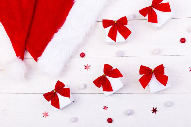 Foto sombrero de navidad y regalos, pequeñas cajas blancas con lazo rojo y dulces sobre fondo blanco de madera.