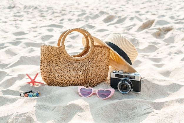 Sombrero de paja, bolso, gafas de sol, coral y cámara sobre arena en playa tropical. Concepto de vacaciones de verano