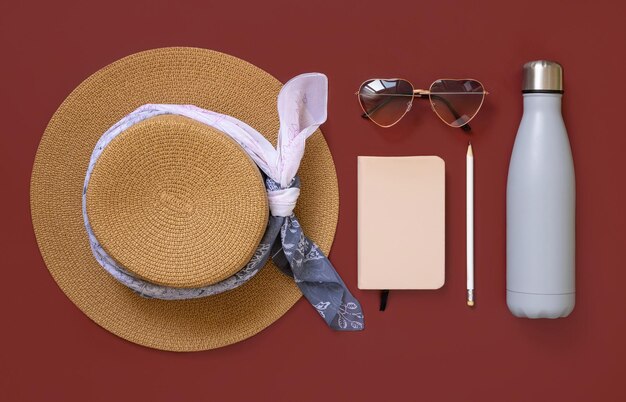 Sombrero de paja, cuaderno, pensil, corazón, gafas de sol y vista superior de botella reutilizable en rojo. Ropa y accesorios de verano femeninos, endecha plana. Libro de texto de tapa dura y maqueta de botella. Concepto de viaje.