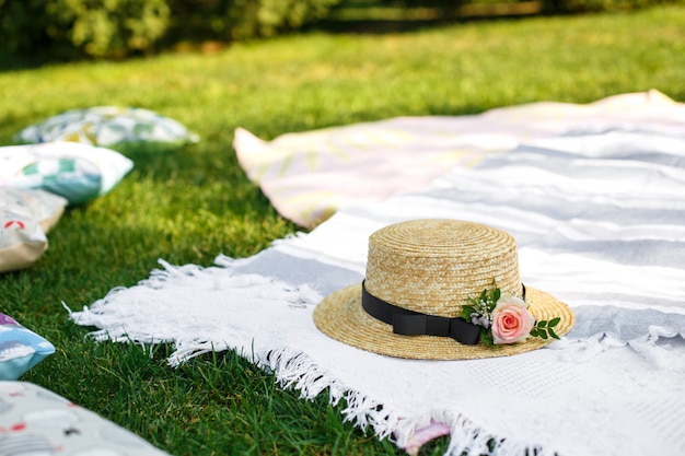 El sombrero de paja con las flores frescas pone en una manta de picnic blanca en el fondo brillante del día de verano del césped verde