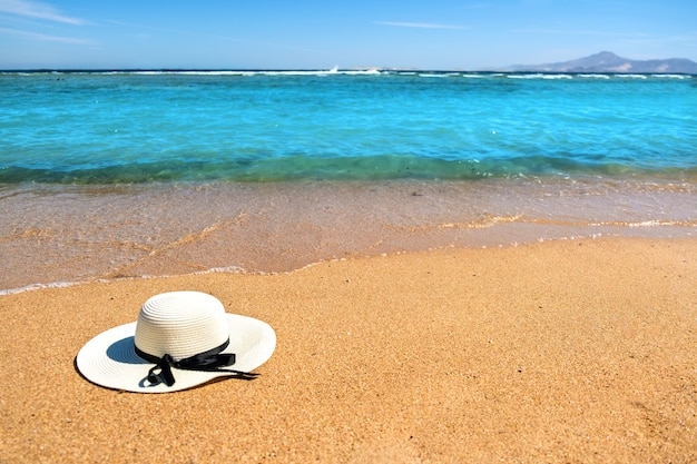 Sombrero de paja de la mujer blanca que pone en la playa tropical de la arena con el agua azul vibrante del océano, día de verano soleado. Concepto de viaje de vacaciones y destino.