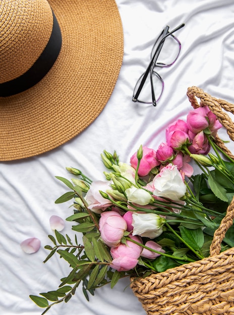 Sombrero de paja y ramo de flores rosas sobre fondo blanco. Vista superior, composición mínima de estilo plano laico. Concepto de vacaciones de verano.