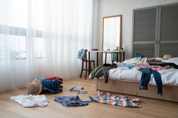 sombrero de paja y ropa colorida en equipaje sobre suelo de madera. vacíe a nadie en la cama blanca desordenada en la maleta de embalaje del dormitorio para viajar al extranjero vacaciones de verano. espejo junto a la ventana del tocador.