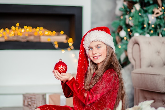 Un sombrero de santa linda chica alegre cerca del árbol de Navidad. Sostiene la bola roja de Navidad.