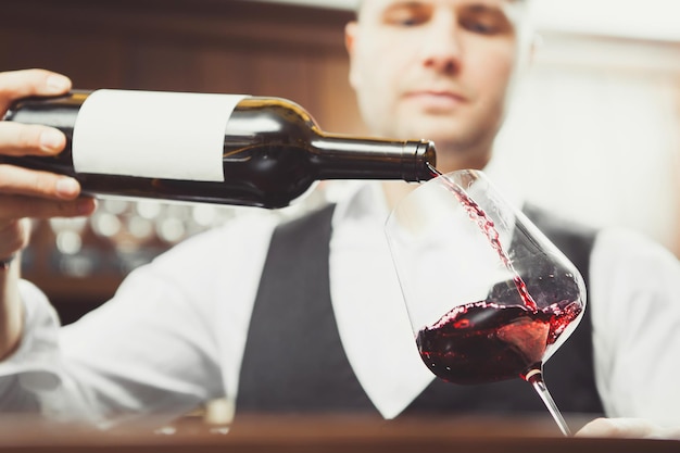 El sommelier de la foto de primer plano vierte vino tinto en un vaso en el fondo del mostrador del bar