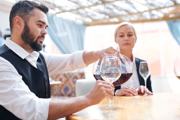 Sommelier o barman joven barbudo vertiendo cabernet rojo en copa de vino antes de degustar