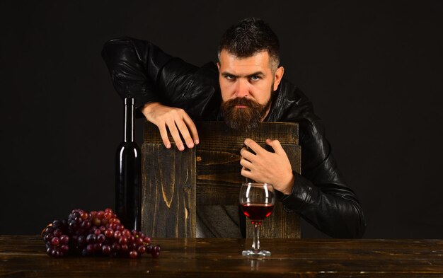 Sommelier se inclina na cadeira de madeira Homem com barba perto de copo de vinho em fundo marrom escuro Degustador com cara séria por garrafa de vinho e uvas escuras Conceito de degustação e degustação