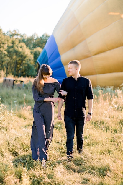 Sommer-Außenaufnahme des freudigen jungen Paares, das während des Sonnenuntergangs im grünen Feld geht und zur Kamera vor dem gelben Heißluftballon aufwirft, der sich auf den Flug vorbereitet