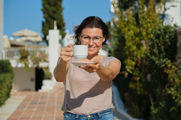 Sommer-Außenporträt der reifen lächelnden Frau, die mit Tasse Kaffee geht