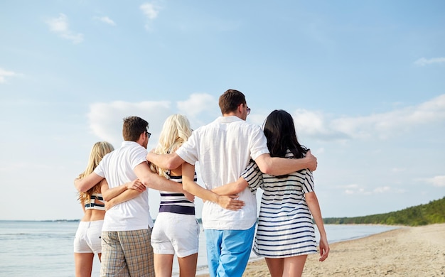 sommer, ferien, meer, tourismus und personenkonzept - gruppe lächelnder freunde, die sich von hinten am strand umarmen und spazieren gehen