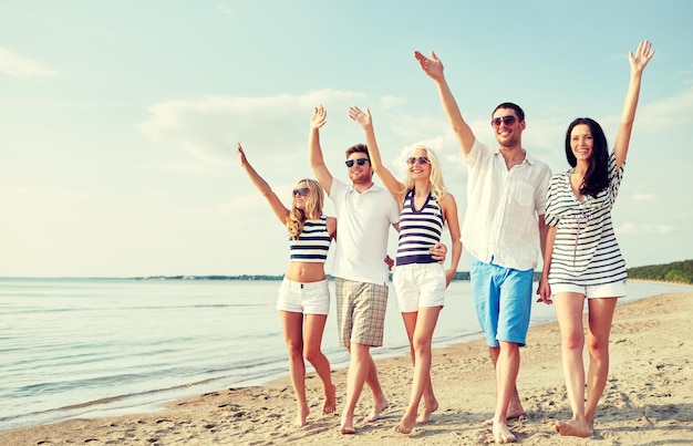 sommer, ferien, meer, tourismus und personenkonzept - gruppe lächelnder freunde mit sonnenbrillen, die am strand spazieren und hände winken