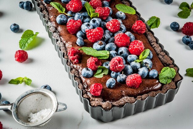 Sommer hausgemachtes Gebäck. Schokoladenkuchentarte mit Schokoladencreme, frische rohe Beeren Heidelbeer Himbeere, dekoriert mit Minze, Puderzucker. Auf weißer Marmortabelle copyspace