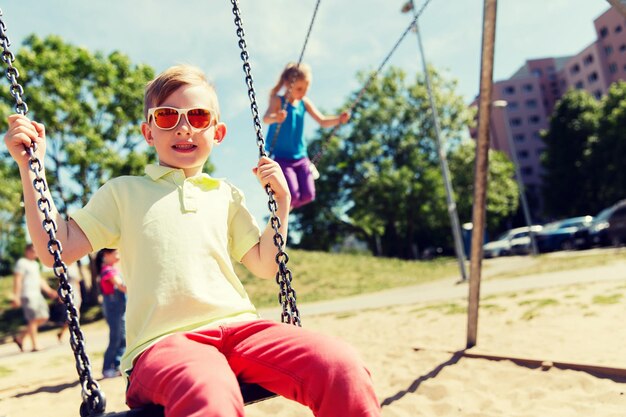 sommer, kindheit, freizeit, freundschaft und menschenkonzept - zwei glückliche kinder, die auf dem kinderspielplatz auf der schaukel schwingen