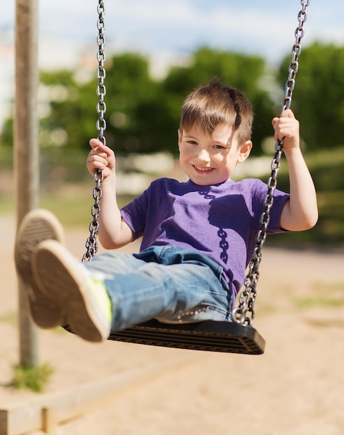 sommer, kindheit, freizeit, freundschaft und personenkonzept - glücklicher kleiner junge, der auf der schaukel auf dem kinderspielplatz schwingt