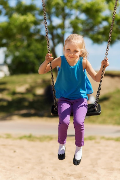 sommer, kindheit, freizeit, freundschaft und personenkonzept - glückliches kleines mädchen, das auf der schaukel auf dem kinderspielplatz schwingt