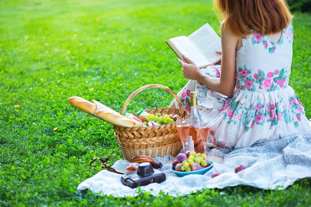 Sommer - Picknick auf der Wiese. Mädchen, das ein Buch liest und in der Nähe eines Picknickkorbs und Baguette, Wein, Gläser, Trauben und Brötchen sitzt