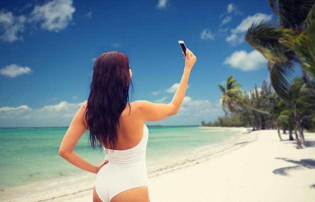 sommer-, reise-, technologie- und personenkonzept - sexy junge frau, die selfie mit smartphone über tropischem strand mit palmenhintergrund macht