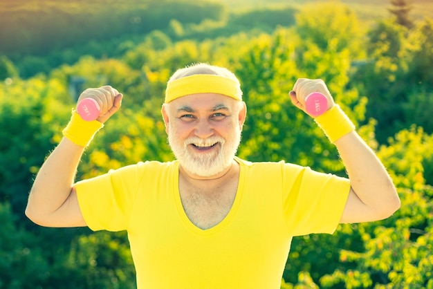 Sommer- und Aktivurlaub Porträt eines älteren Mannes, der eine Hantel hält Glücklicher älterer Sportler, der mit anhebender Hantel auf grünem Parkhintergrund trainiert