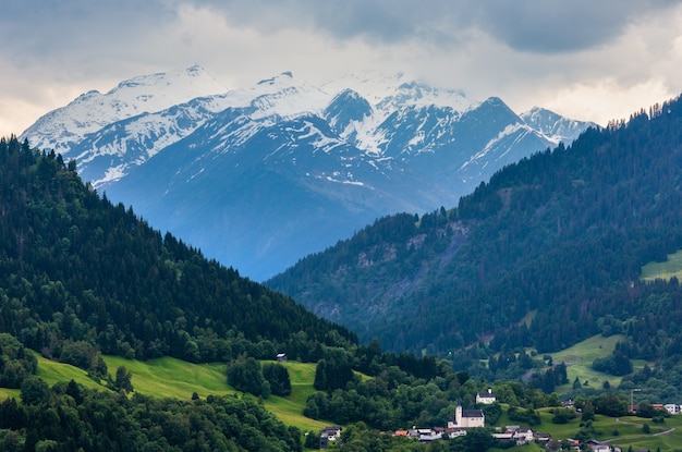 Sommeralpengebirgslandschaft mit Dorf, Tannenwald am Hang und schneebedeckten Felsgipfeln im fernen Österreich.