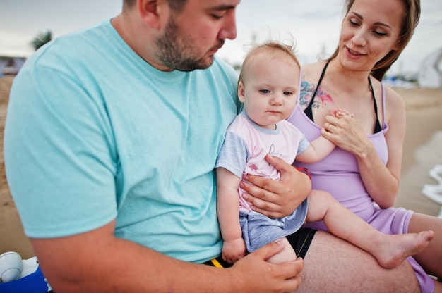 Sommerferien. Eltern und Menschen im Freien mit Kindern. Schöne Familienferien. Vater, schwangere Mutter, kleine Tochter, die auf der Sonnenliege am Meeressandstrand sitzt.