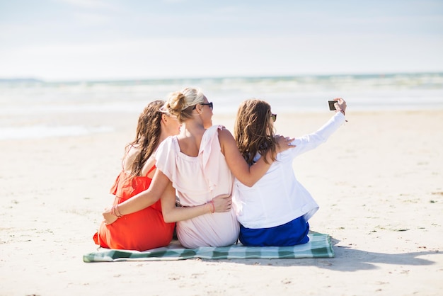 Foto sommerferien, ferien, reisen, technologie und personenkonzept - gruppe lächelnder junger frauen, die selfie mit smartphone am strand machen