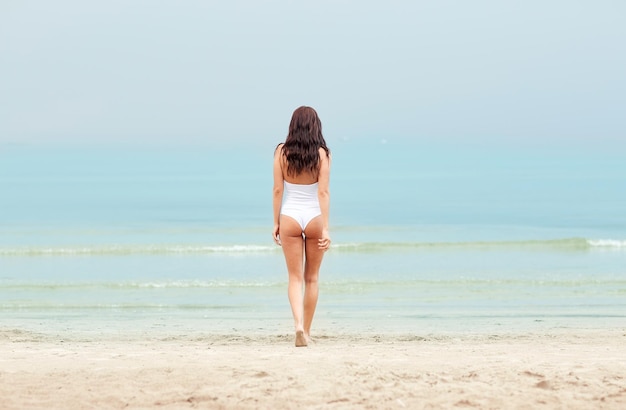 sommerferien, tourismus, reise, urlaub und personenkonzept - junge frau im badeanzug, die von hinten am strand spazieren geht