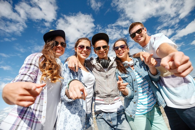 sommerferien und teenagerkonzept - gruppe lächelnder teenager mit sonnenbrillen, die draußen hängen und mit dem finger auf sie zeigen