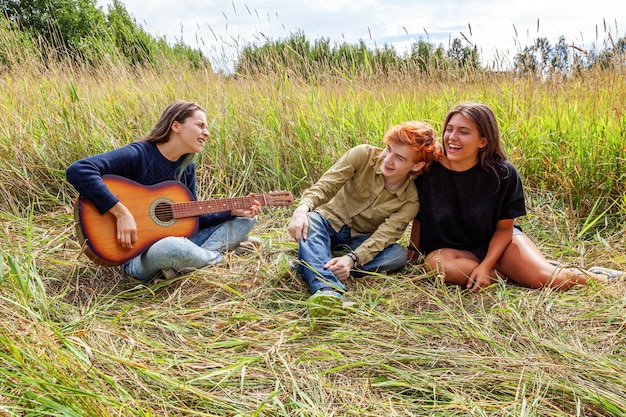 Sommerferien Urlaub Musik glückliche Menschen Konzept. Gruppe von drei Freunden, Jungen und zwei Mädchen mit Gitarrensingen, die gemeinsam Spaß im Freien haben. Picknick mit Freunden auf einem Roadtrip in der Natur.