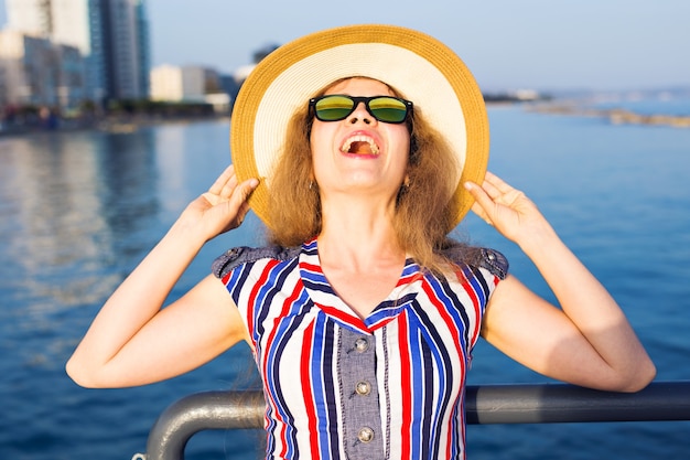 Sommerferien, Urlaub, Reisen und Menschen-Konzept - lächelnde lachende junge Frau mit Sonnenbrille und Hut am Strand über dem Meer.