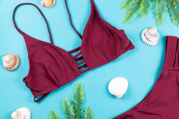 Sommerferienhintergrund. Tropisches Sommerkonzept mit rotem Bikini, Blättern und Muscheln auf blauem Hintergrund.