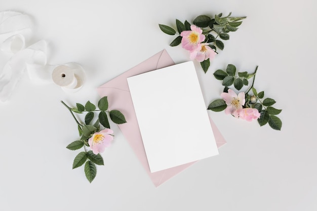 Sommerhochzeit Briefpapier Mockup Szene mit Seidenband Blanko Grußkarte Einladung rosa Umschlag und blühende Hunderosenblüten isoliert auf weißem Tischhintergrund Flache Ansicht von oben