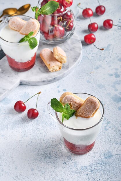 Sommerkirschblätterteig mit Savoiardi-Keksen und Frischkäse im Glas auf hellgrauem Hintergrund. Traditioneller Tiramisu-Kuchen mit frischen Beeren. Selektiver Fokus.