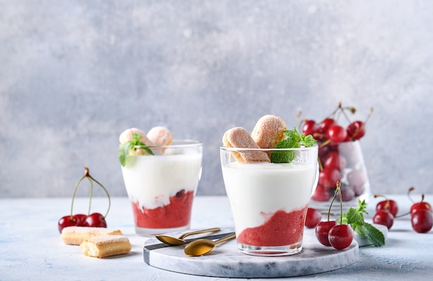 Sommerkirschenblätterteig mit Savoiardi-Keksen und Frischkäse im Glas auf hellgrauem Hintergrund. Traditioneller Tiramisu-Kuchen mit frischen Beeren. Selektiver Fokus.