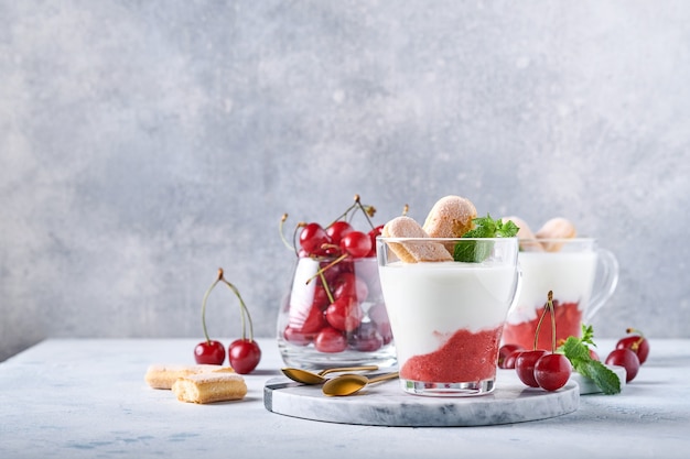 Sommerkirschenblätterteig mit Savoiardi-Keksen und Frischkäse im Glas auf hellgrauem Hintergrund. Traditioneller Tiramisu-Kuchen mit frischen Beeren. Selektiver Fokus.