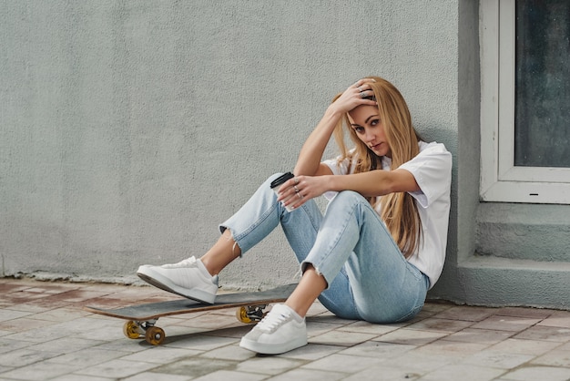 Sommerlebensstil des modernen recht jungen Mädchens, das nahe bei einem Skateboard sitzt.