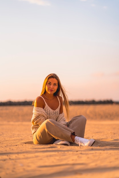 Sommerlebensstil, eine junge Blondine mit glattem Haar, in einem kleinen Wollpullover und einer Cordhose am Strand sitzend. Im ernsthaften Sand sitzen