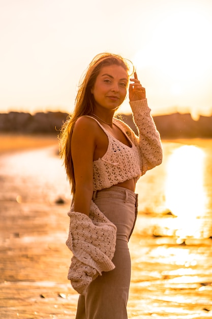 Sommerlicher Lebensstil, eine junge Blondine mit glattem Haar, die einen kleinen Wollpullover und eine Cordhose am Strand trägt. Die Sonne bei Sonnenuntergang genießen