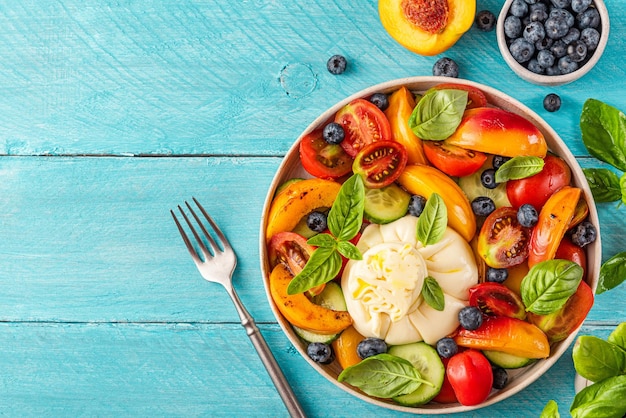 Sommerlicher Obst- und Gemüsesalat mit Burrata-Käse und Basilikum in einem Teller auf blauem Holztisch für ein leckeres Mittagessen