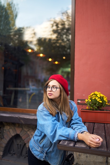Sommerliches sonniges Lebensstilmodeporträt der jungen stilvollen Hipsterfrau, die auf Straße geht und niedliches trendiges Outfit mit rotem Hut trägt