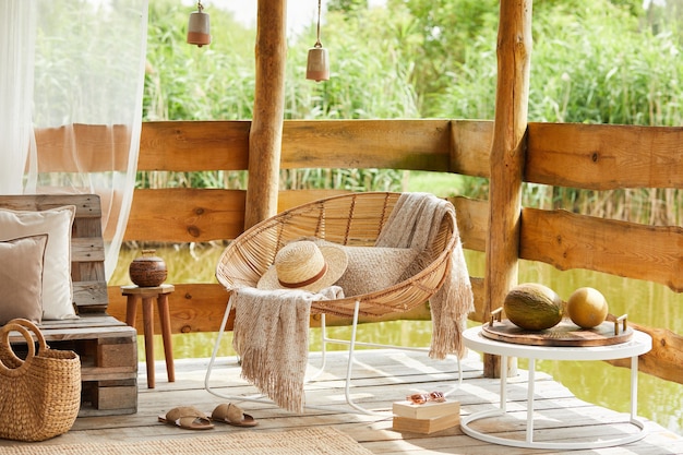 Sommerpavillon am See mit stilvollem Rattansessel, Couchtisch, Sofa, Kissen, Plaid und eleganten Accessoires in modernem Dekor. Sommergefühl. Beruhige dich.