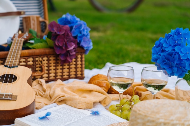 Sommerpicknick am sonnigen Tag mit Brot, Obst, Bouquet Hortensienblüten, Gläsern Wein, Strohhut, Buch und Ukulele.